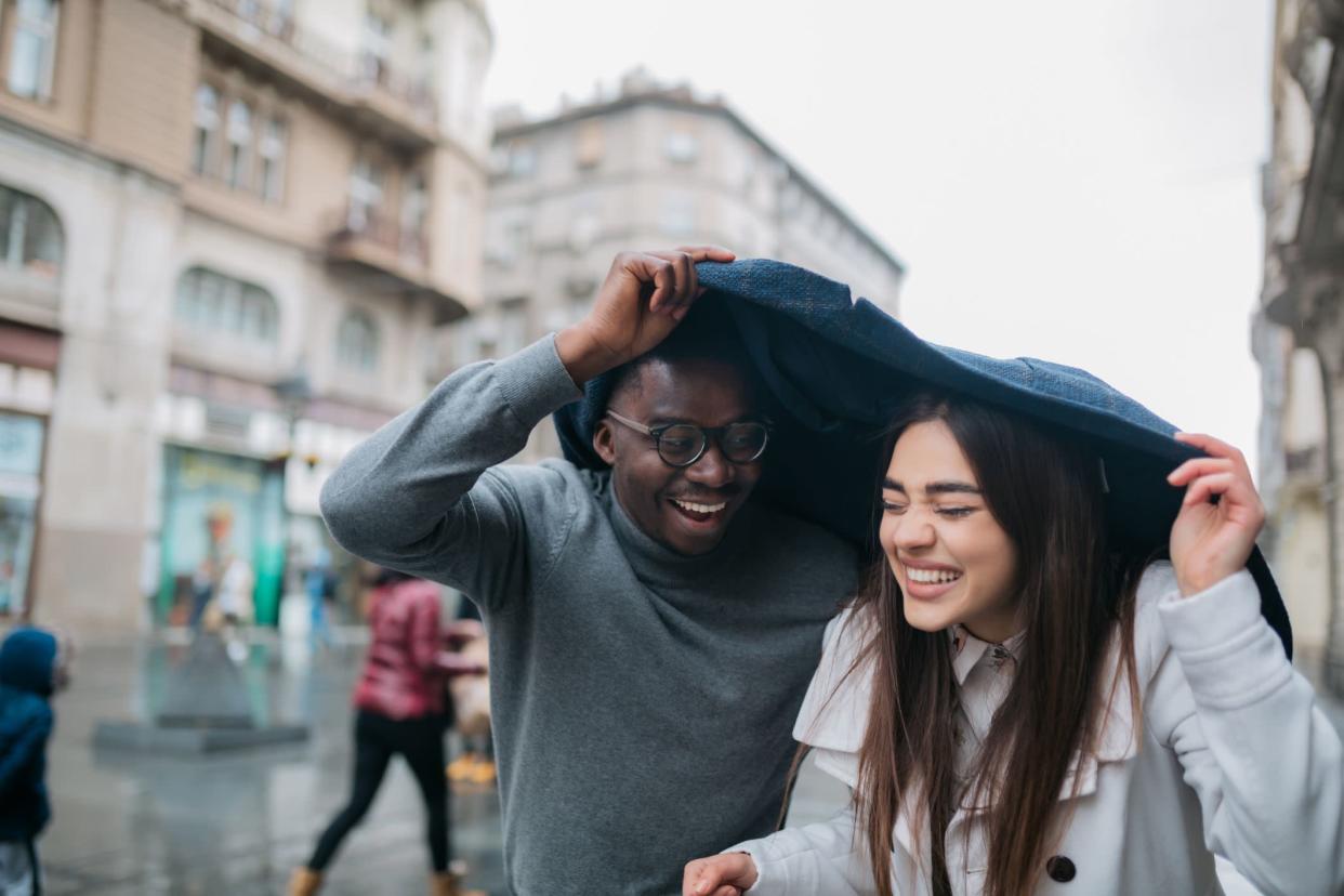 Young diverse couple having fun together, walking around the city on a rainy day covering themselves with a jacket to protect themselves from the rain, smiling