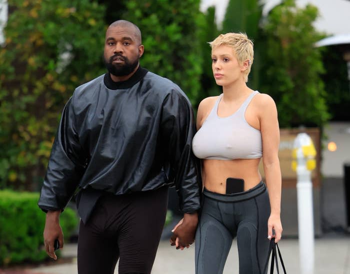 Ye in a black jacket and pants holding hands with Bianca, who's in a gray sports crop top and leggings, both walking
