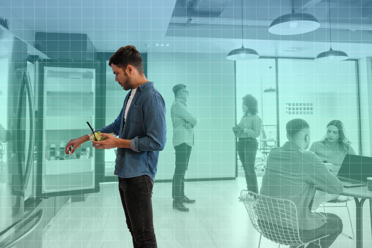 a photo of a man in an office kitchen looking in the refrigerator with his coworkers working behind him