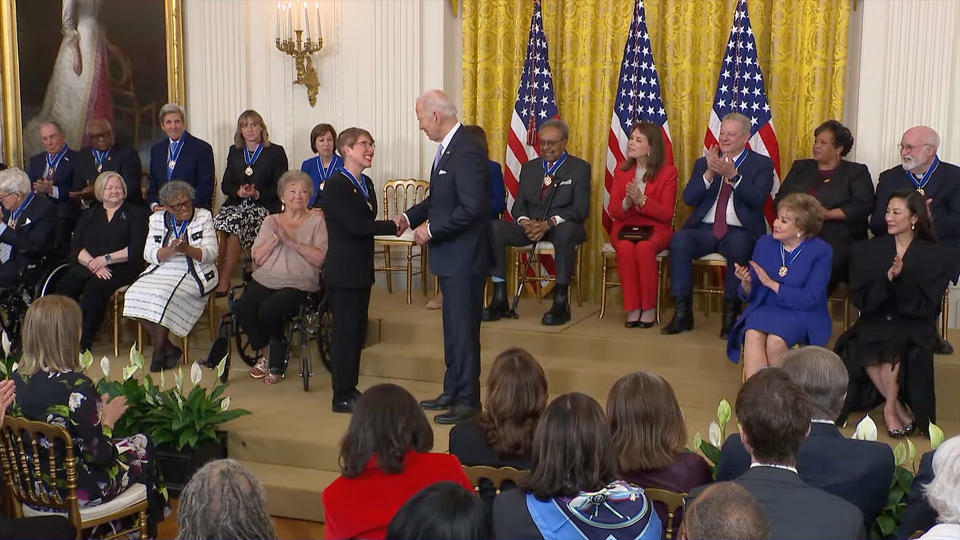 A man in a blue suit shakes hands with a woman in a navy blue suit in a crowded room with multiple American flags