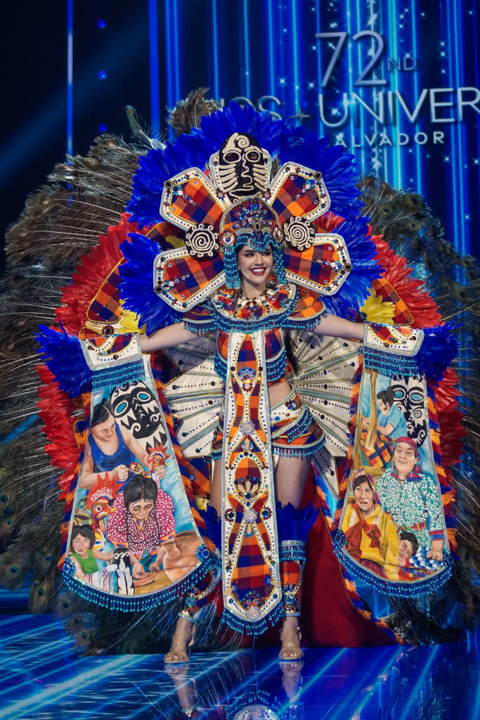 She's wearing an ornate two-piece outfit and ornate headdress with a train that has illustrations of Native scenes