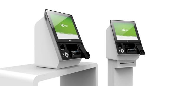 Two kiosks with NCR logos on the screen, sitting on white pedestal and desk.