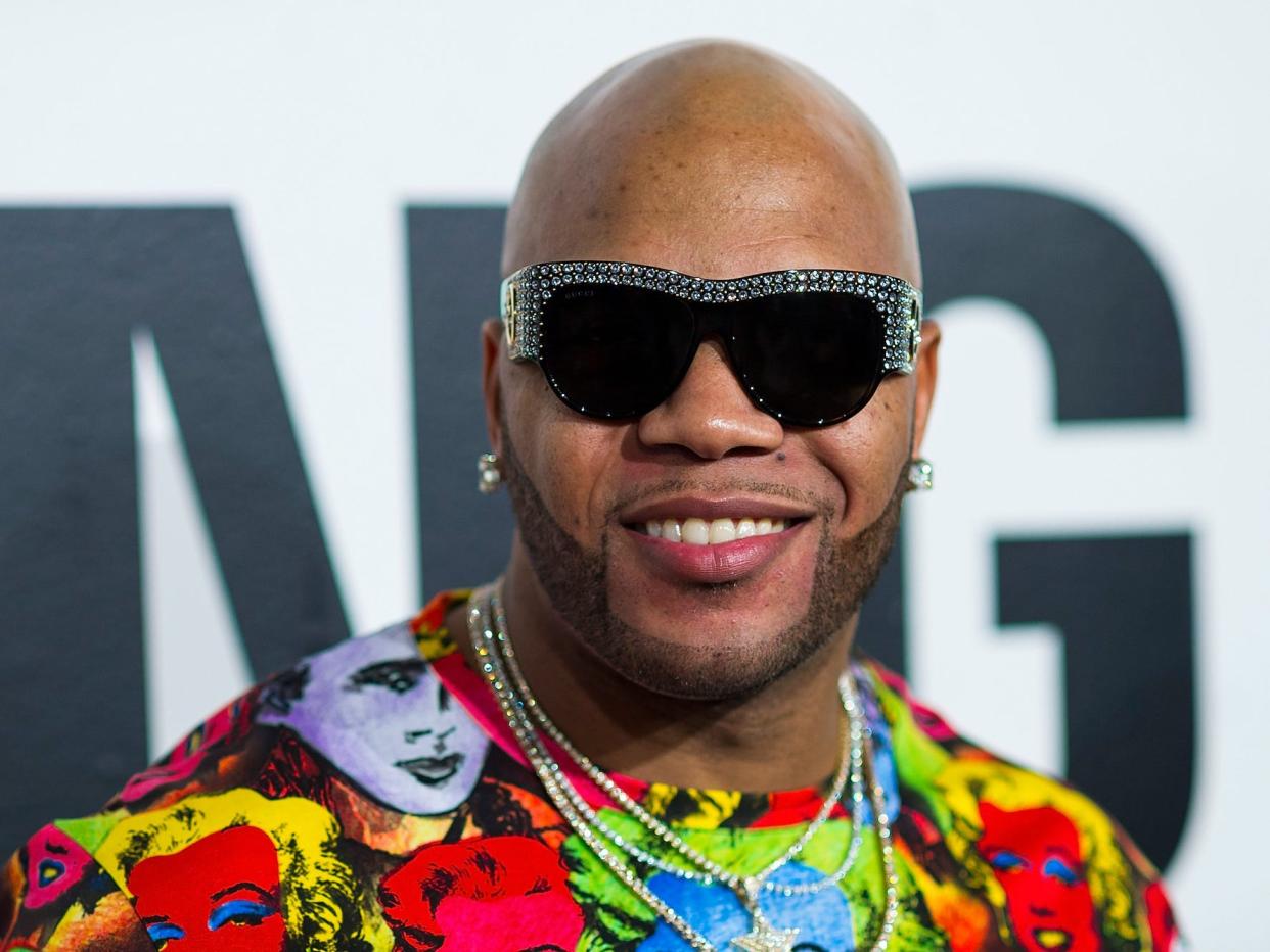 Flo Rida posing for photographers at the Samsung Charity Gala while wearing blacked out sunglasses and a bright multicoloured shirt.