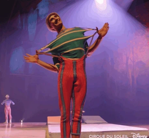 A Cirque du Soleil performer is stretching his shirt while wearing striped pants and a beanie during a stage act