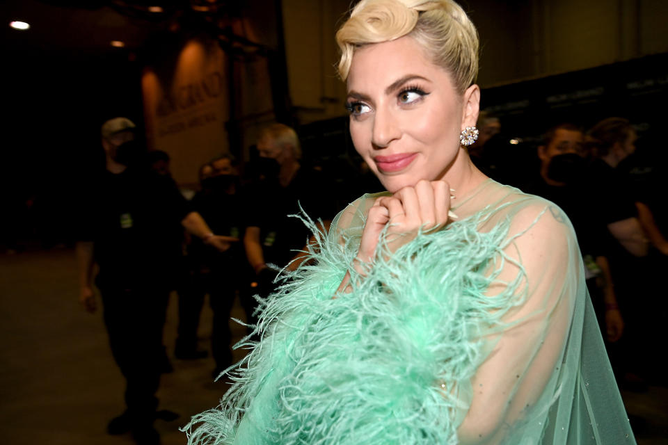 Lady Gaga in a green feathered outfit, smiling with hands under chin, at an event