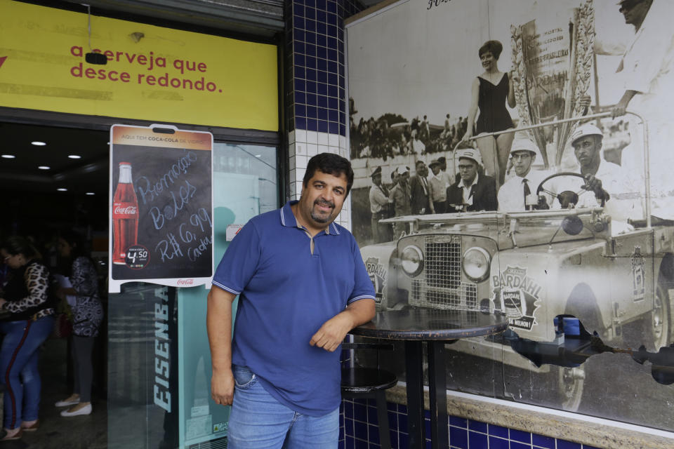 In this Nov. 7, 2019 photo, Marcos Resenti, owner of a bakery located next to the Interlagos racetrack, poses for a photo in front of an old photo showing a race at Interlagos, in Sao Paulo, Brazil. Resenti said he was was shocked when he heard President Jair Bolsonaro announced in June there was a 99% chance F1 would move to Rio de Janeiro. (AP Photo/Nelson Antoine)