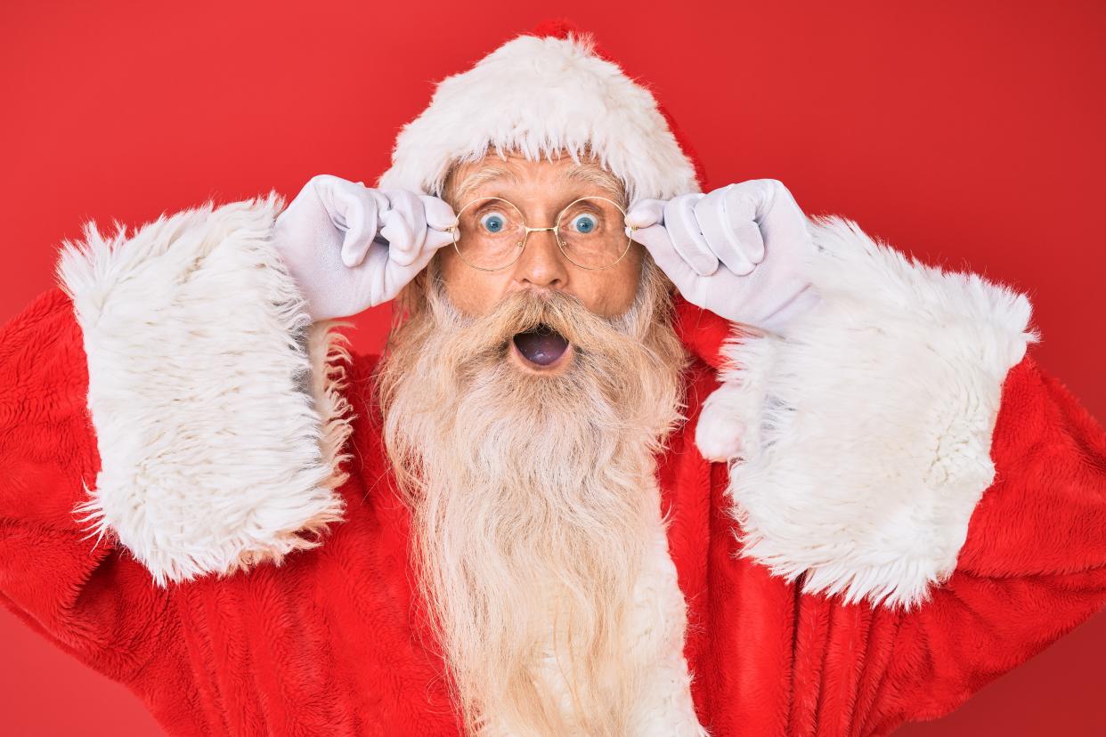  Father Christmas - an old man with white hair and a long white beard wearing a red fur-trimmed hat and coat and circular glasses - holds his glasses and opens his mouth in a shocked expression. 
