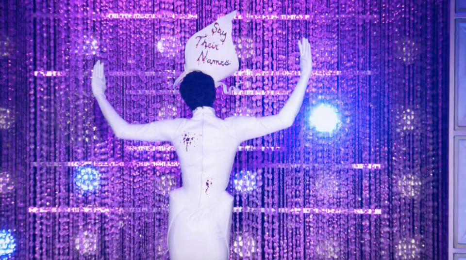 A drag queen in a floor length white dress with a fascinator hat inscribed, "Say Their Names."