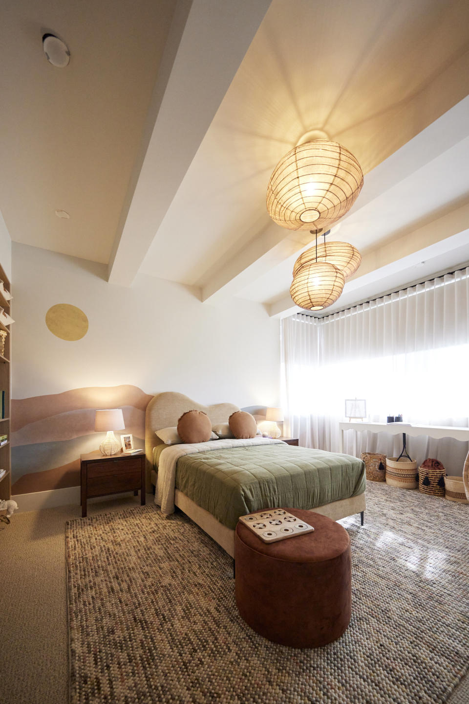 A wide shot of the bedroom with a double bed, an ottoman in front of it, on a textured rug with material light fixtures hanging above it. 