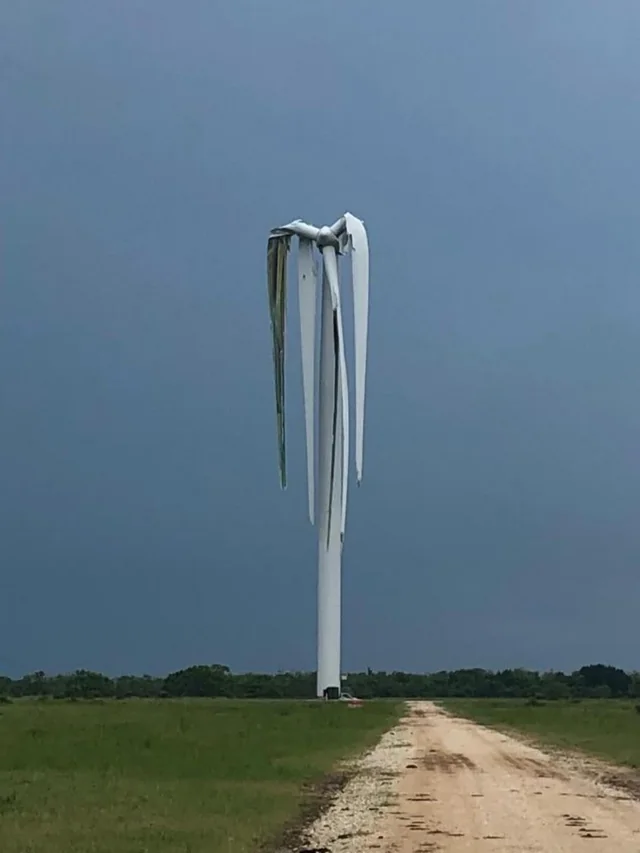 A broken wind turbine stands tall in a field, with its blades bent and twisted downward. A dirt path leads towards it under a dark, cloudy sky