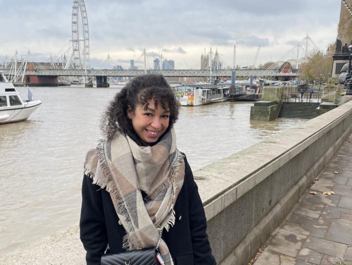 Amber Lucas, wearing a black coat and a scarf, poses in London near the River Thames while visiting the city with a date