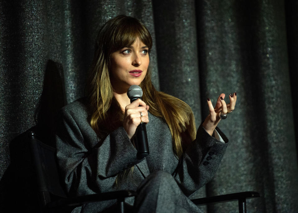 Photo of Dakota Johnson in a gray suit, sitting in a chair, gesturing with her hand, and talking into a microphone
