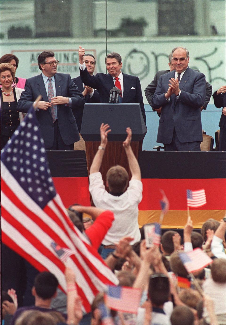 <p>Ein Zuschauer springt in die Höhe, nachdem US-Präsident Ronald Reagan vor dem Brandenburger Tor in Westberlin eine Rede gehalten hat. Neben ihm stehen der damalige BRD-Kanzler Helmut Kohl und der Präsident des Deutschen Bundestages Philipp Jenninger. Reagan hatte am 12. Juni 1987 dazu aufgerufen, die Mauer niederzureißen – und ging mit den Worten “Mr. Gorbatschow, tear down this wall” in die Geschichte ein. (Bild: AP Photo/J. Scott Applewhite) </p>