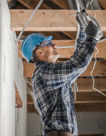 A man in a flannel shirt and a blue hard hat installs wires in a room with exposed beams.