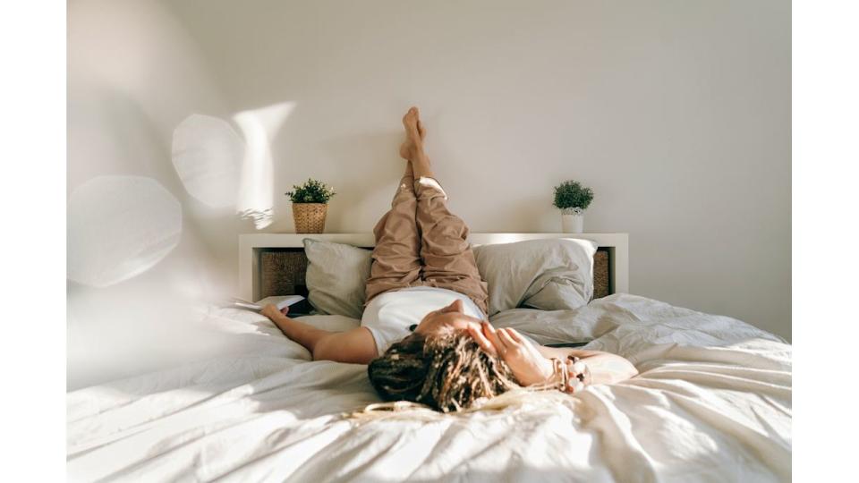 Woman in home clothes book lying on the bed reading. Work at home. Portrait of a dreadlocks braid woman legs up, inverted pose, swelling prevention varicose veins. High angle view