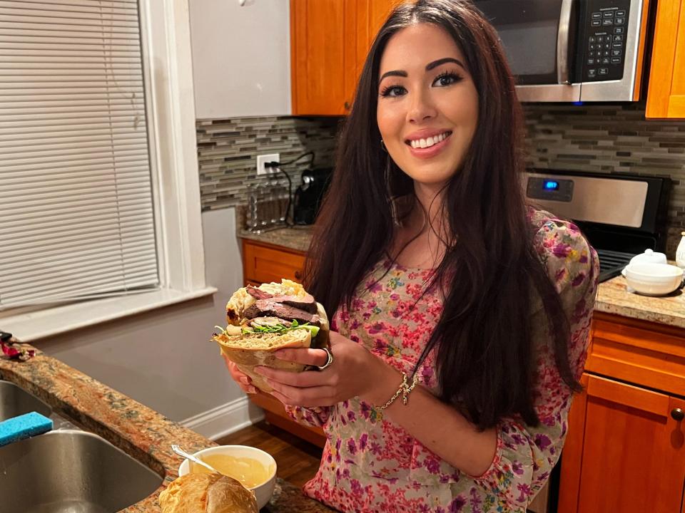 Lara smiles and holds up half of the assembled steak sandwich in her kitchen.