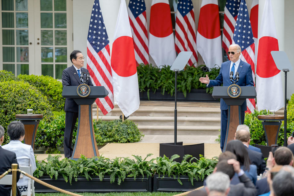 two men in suits stand lecturing outside in front of four Japanese flags and four American flags