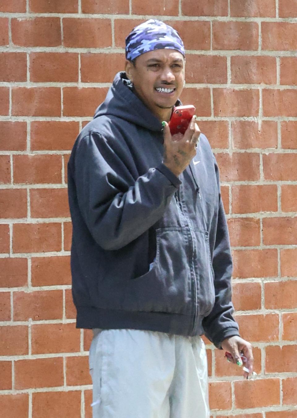 Alexander Edwards stands in front of a brick wall, holding a red phone and smiling. They are wearing a bandana, a hooded jacket, and light-colored pants