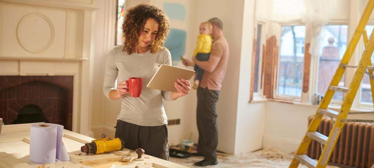 A-woman-looks-at-a-tablet-while-a-man-holds-a-baby-in-the-background-of-a-house-during-repairs.