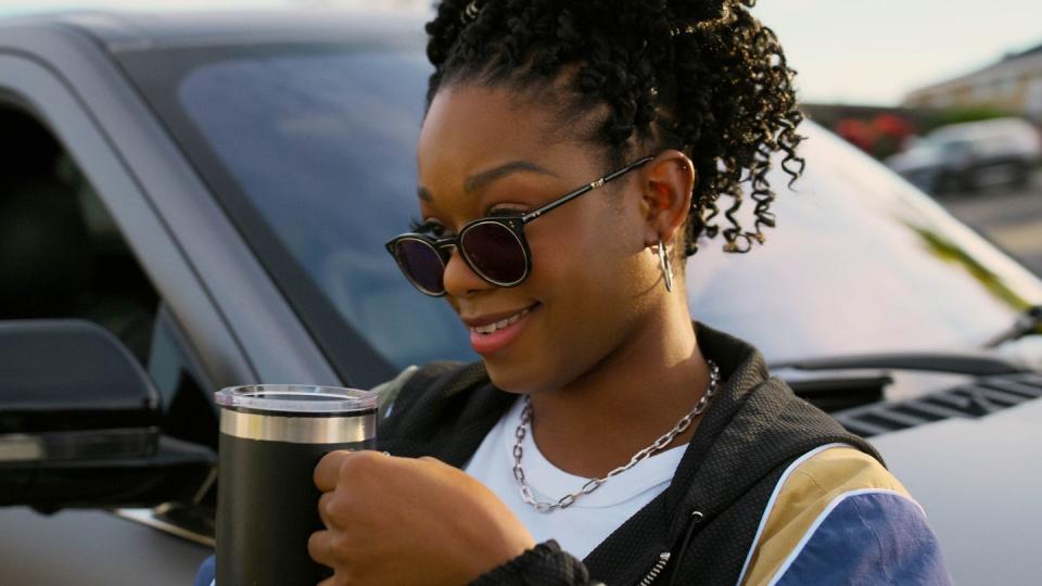 izzy's character stands beside a lincoln suv drinking a cup of coffee, wearing sunglasses