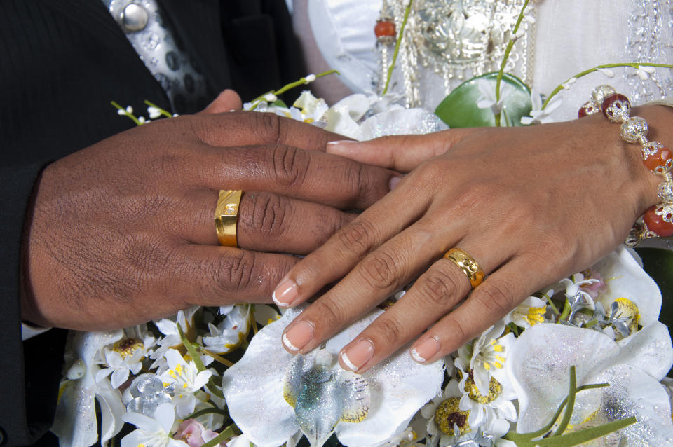 Hands of a bride and groom with wedding rings shown on a bouquet of white flowers, bride with manicured nails