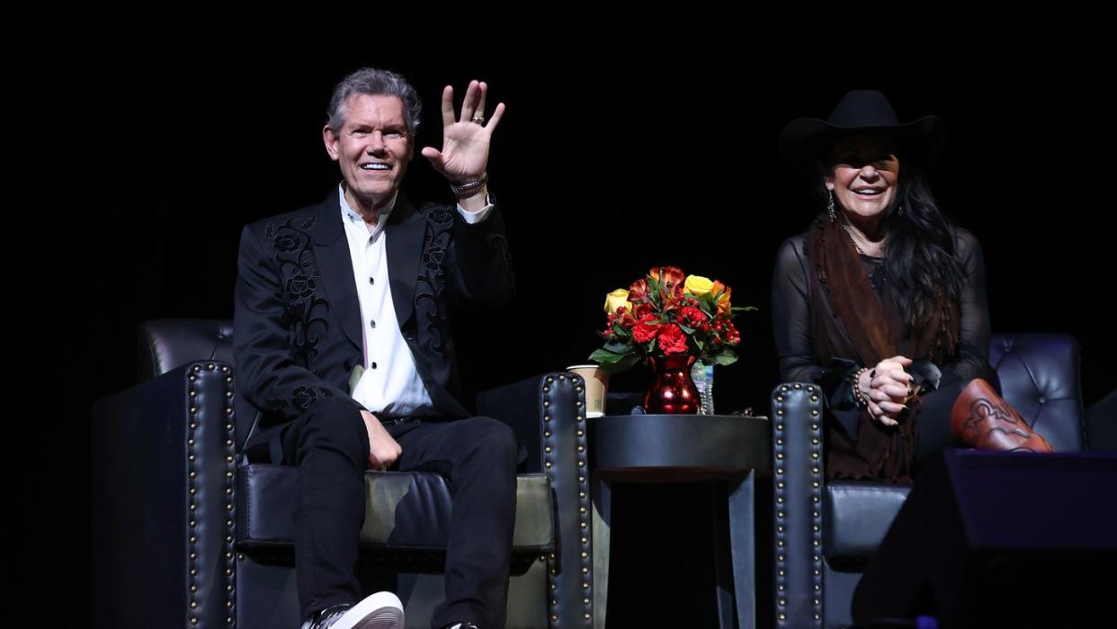 randy travis sitting in a chair smiling and waving with his left hand