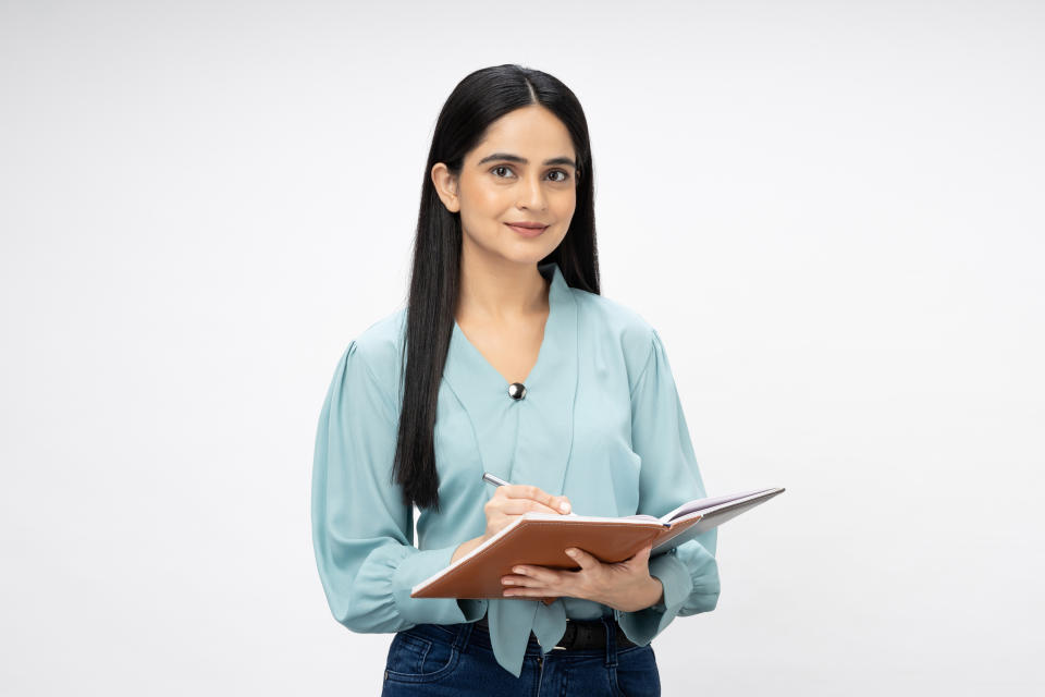 A woman with long, straight hair wearing a professional blouse holds a notebook and pen, smiling slightly while looking at the camera