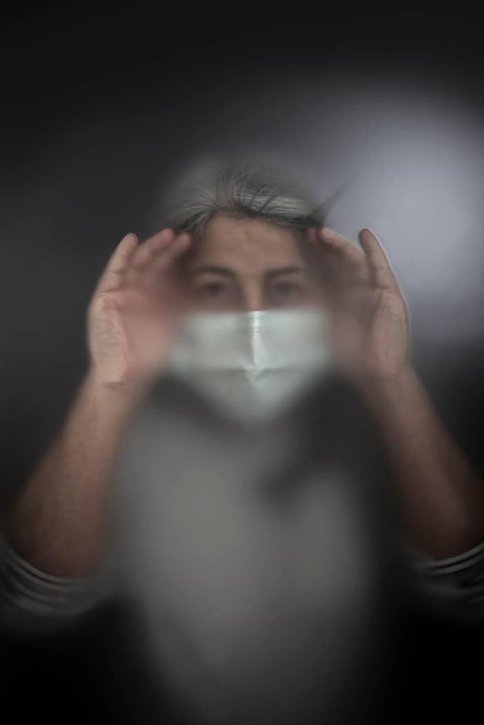 Person with mask looking through a foggy transparent surface with hands pressed against it
