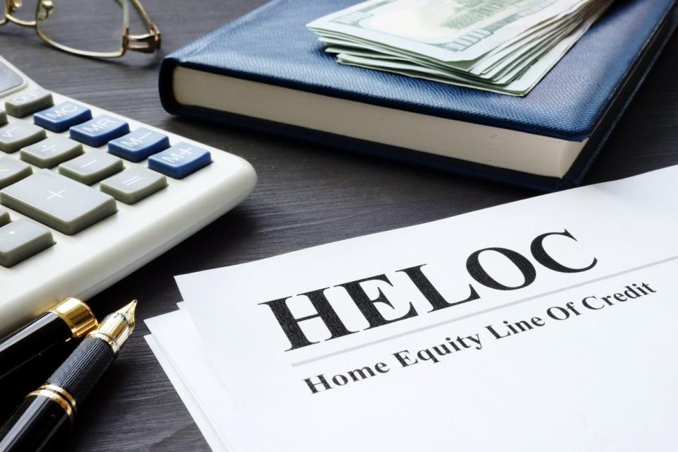 A form with the words 'HELOC home equity line of credit' on it sits on a table with money, a book, glasses, a calculator, and a fountain pen.