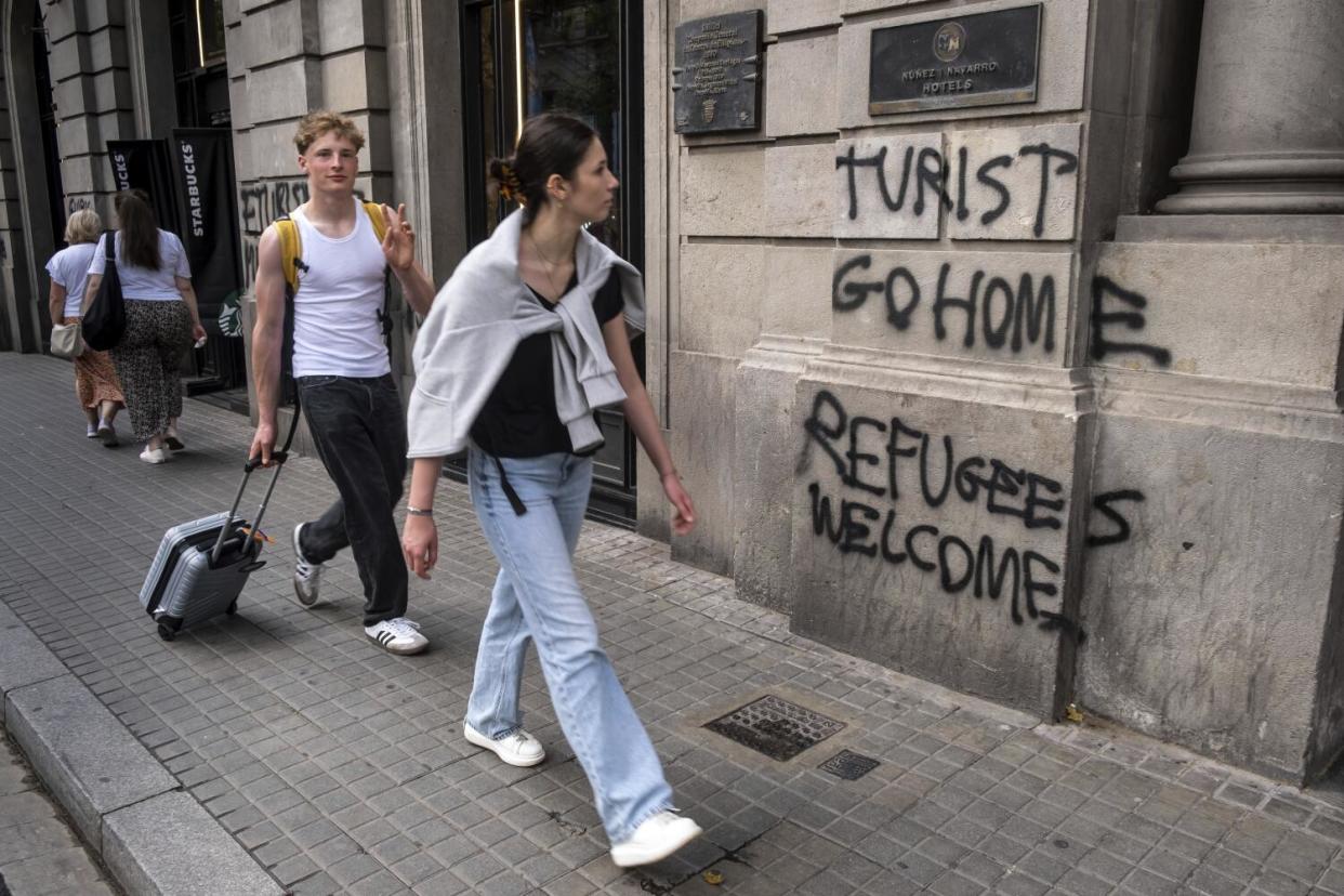 Two pedestrians, one pulling a suitcase, pass a building with black graffiti reading "Turist go home" and "Refugees welcome"