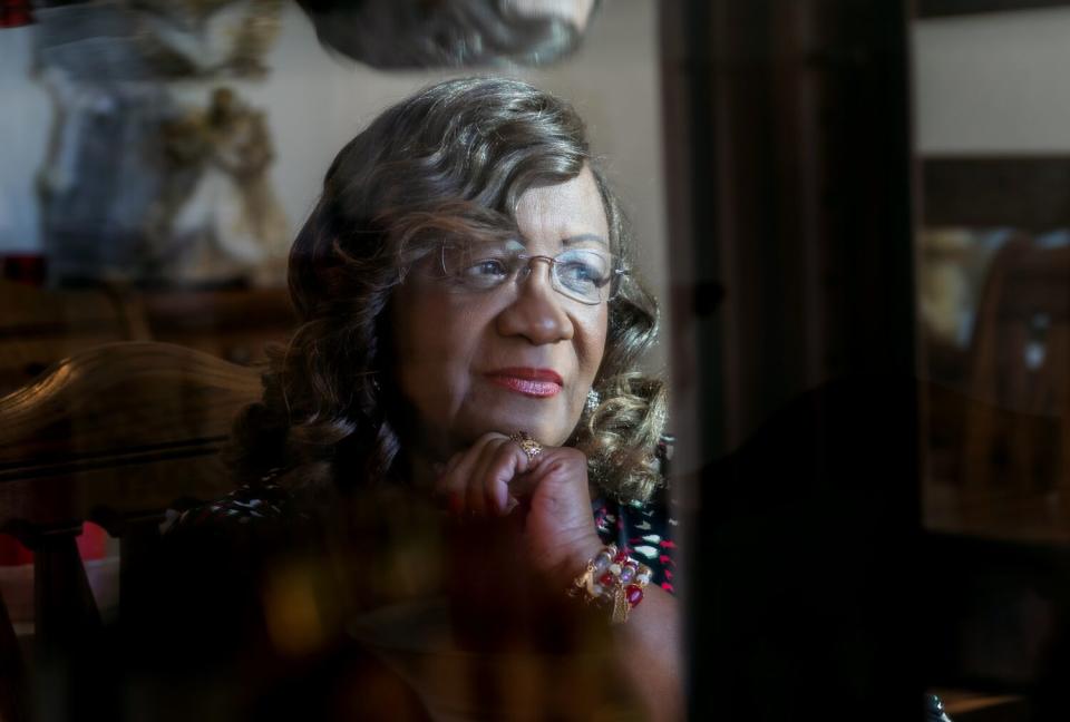 Pearl Devers, partially framed through furniture, sitting near window light, resting her chin on her fist, looking pensive