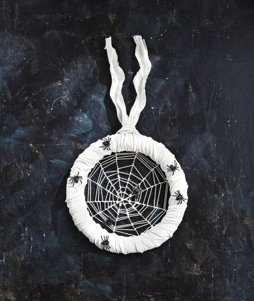 <p>This wreath will definitely give your guests the creepy crawlies. Enter if you dare!</p><p><strong>Make the Spider Wreath: </strong>Tie six pieces of white string across a 14-inch foam wreath form, making sure to loop each one at the midway point of the first piece attached to create a central point. (This is the base of the web and should have 12 "spokes.") Tie a long piece of string to the center point; weave and loop from the center out to create the web. If you run out of string, tie another piece to the end and continue weaving. When you reach the wreath form, tie off at your ending point. Move the twine up and down to create uneven gaps in the web. Wrap the wreath form with white burlap ribbon and attach faux spiders with hot-glue. Loop a piece of white burlap ribbon around the form to hang. <br></p>