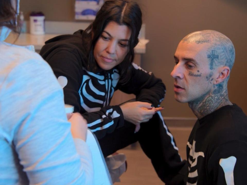 kourtney kardashian sits on a medical examination table, while travis barker kneels near her feet. both are looking at a nurse, whose back is visible in the photo