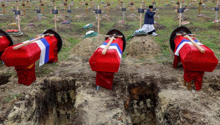 Four coffins covered in red sheets beside open graves, with lines of grave markings behind them