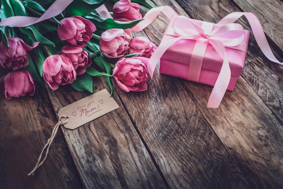 Vintage look of a pink gift box and tulips bouquet and a brown craft tag with the text "Love you Mom" shot on rustic wooden table.