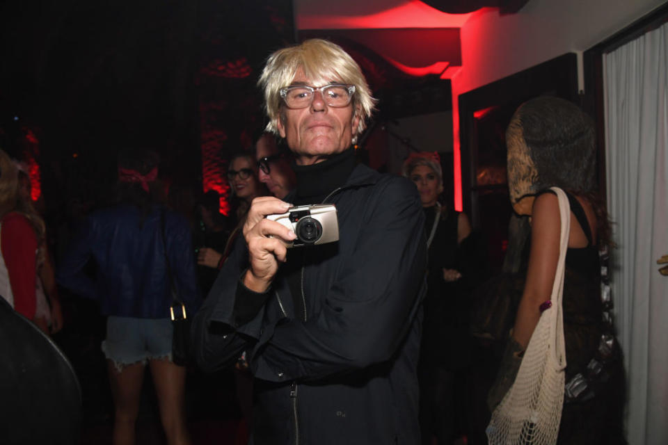 Harry Hamlin dressed as Andy Warhol in a crowd is holding a camera and looking directly at it. He has white hair and is wearing glasses and dark clothing