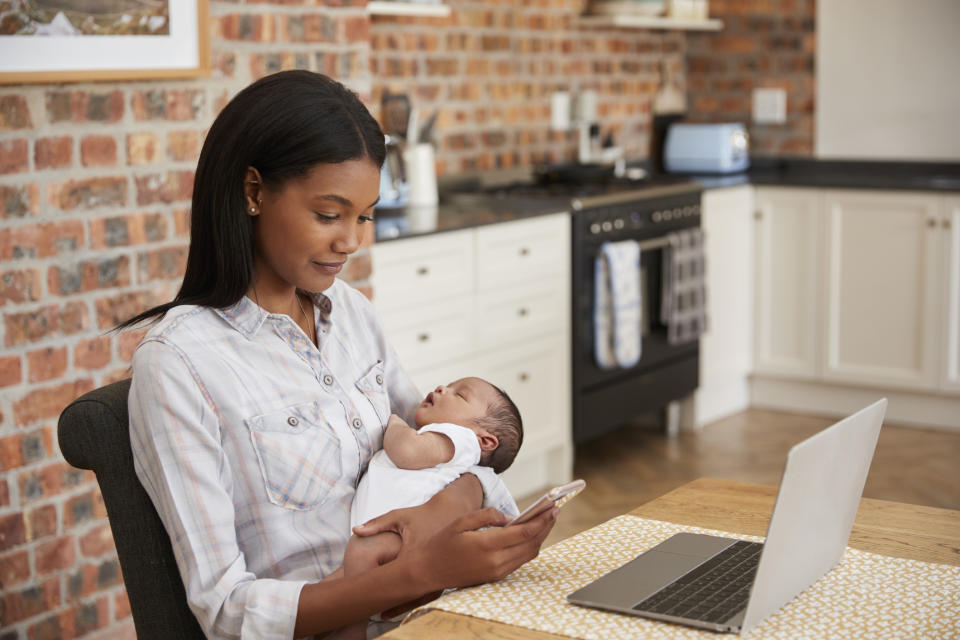 Woman at laptop sitting in a kitchen and checking phone while holding baby.