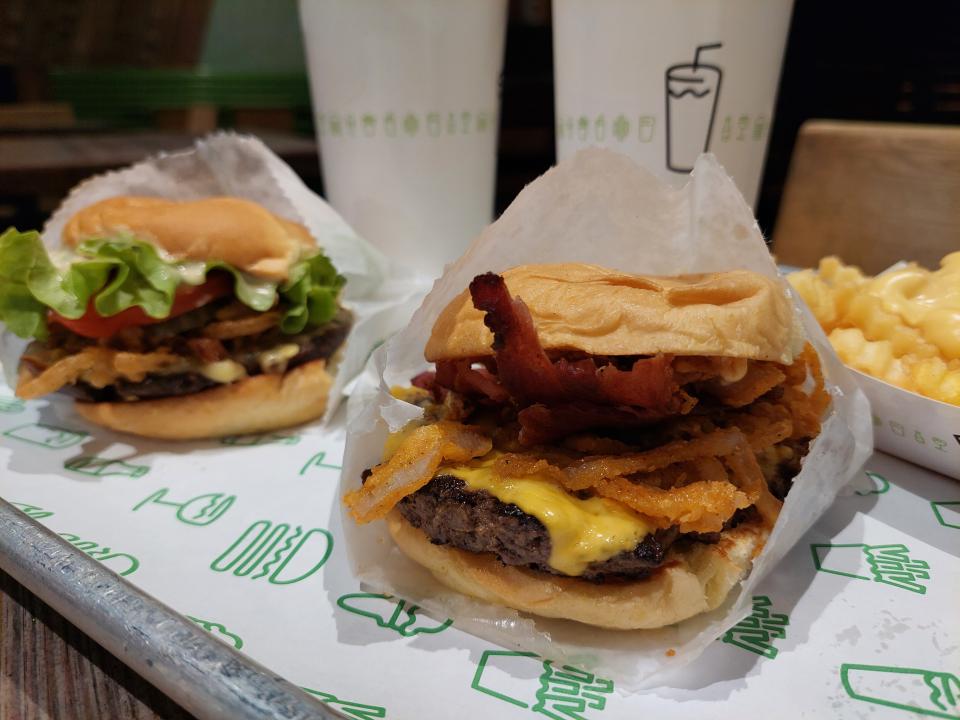 A selection of food from Shake Shack, displayed on a tray in a restaurant: a crispy shallot burger, a ShackMeister with bacon, Shack sauce, cheesy fries, two drinks
