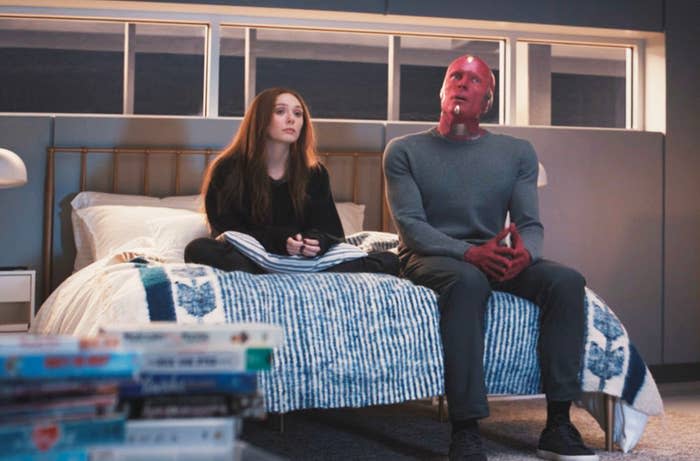 Elizabeth Olsen and Paul Bettany as their characters sit on a bed with a striped blanket in a modern room, looking forward with neutral expressions