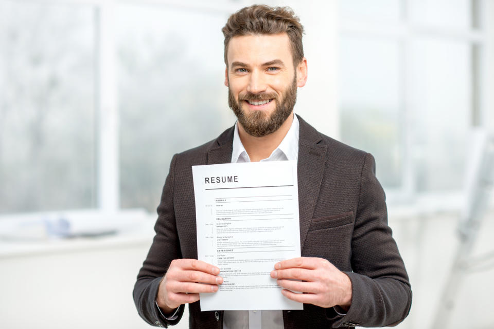A man in a suit holding a resume towards the camera, smiling