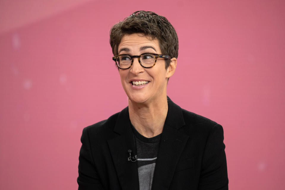 I don't know who this is. A person with short hair, wearing glasses, a blazer, and a t-shirt is smiling against a plain backdrop
