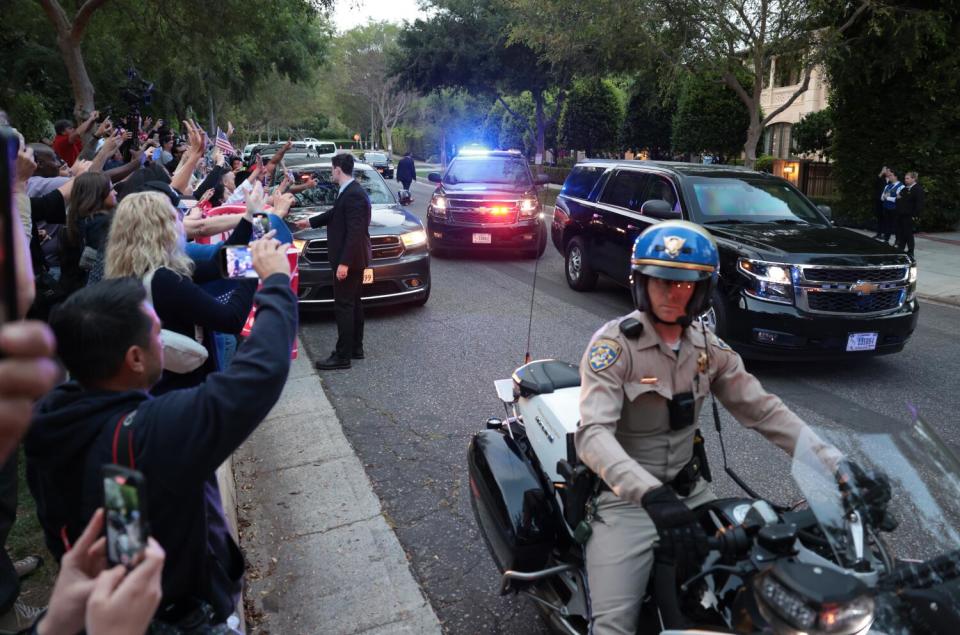 Vehicles drive down a street while onlookers wave.