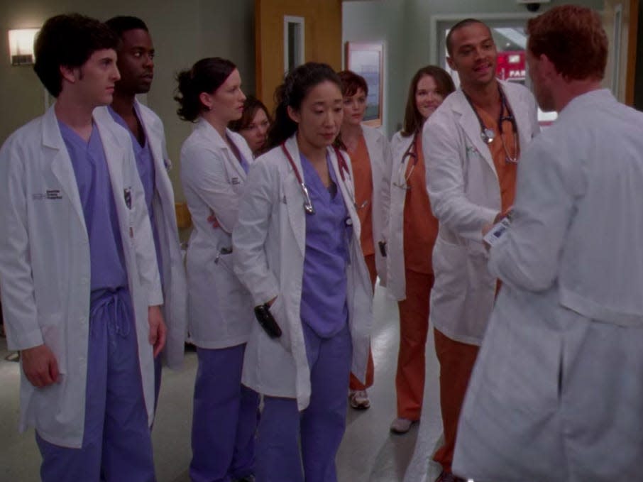 The cast of greys anatomy standing in the lobby wearing scrubs and white coats