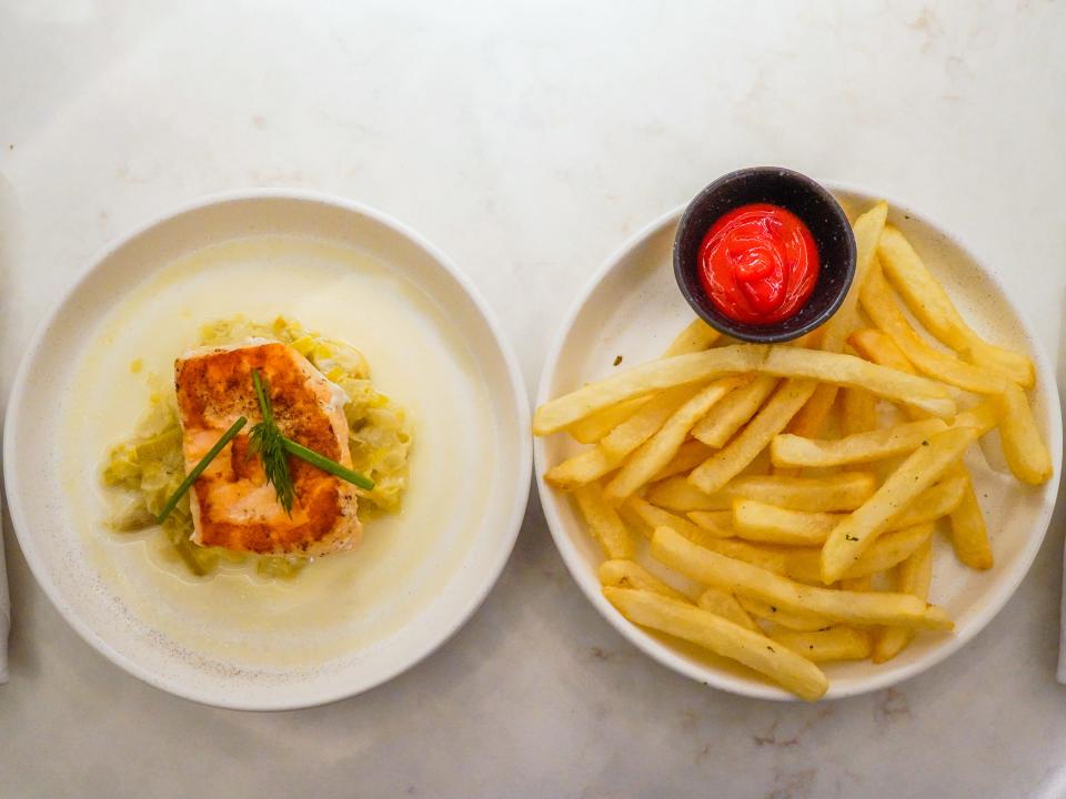 Two white plates side-by-side on a white table. On the left plate is a salmon dish, and on the right plate is fries and ketchup