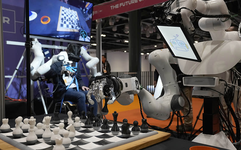 A robot in the foreground makes a chess move while a human in the background controls the robot remotely