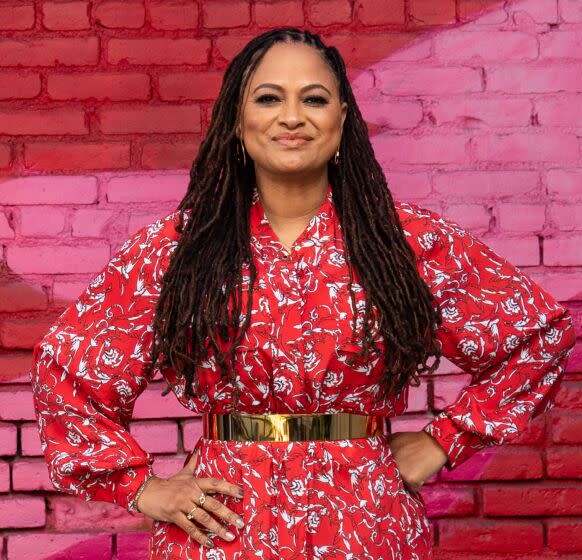 A woman with long dark braids poses in a bright red dress in front of a pink and red background