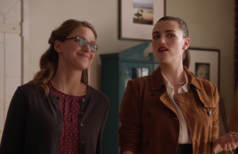 Kara Danvers, wearing a red polka dot shirt under a brown cardigan, smiles as Lena Luthor, wearing a white blouse under a brown jacket, has a skeptical look on her face