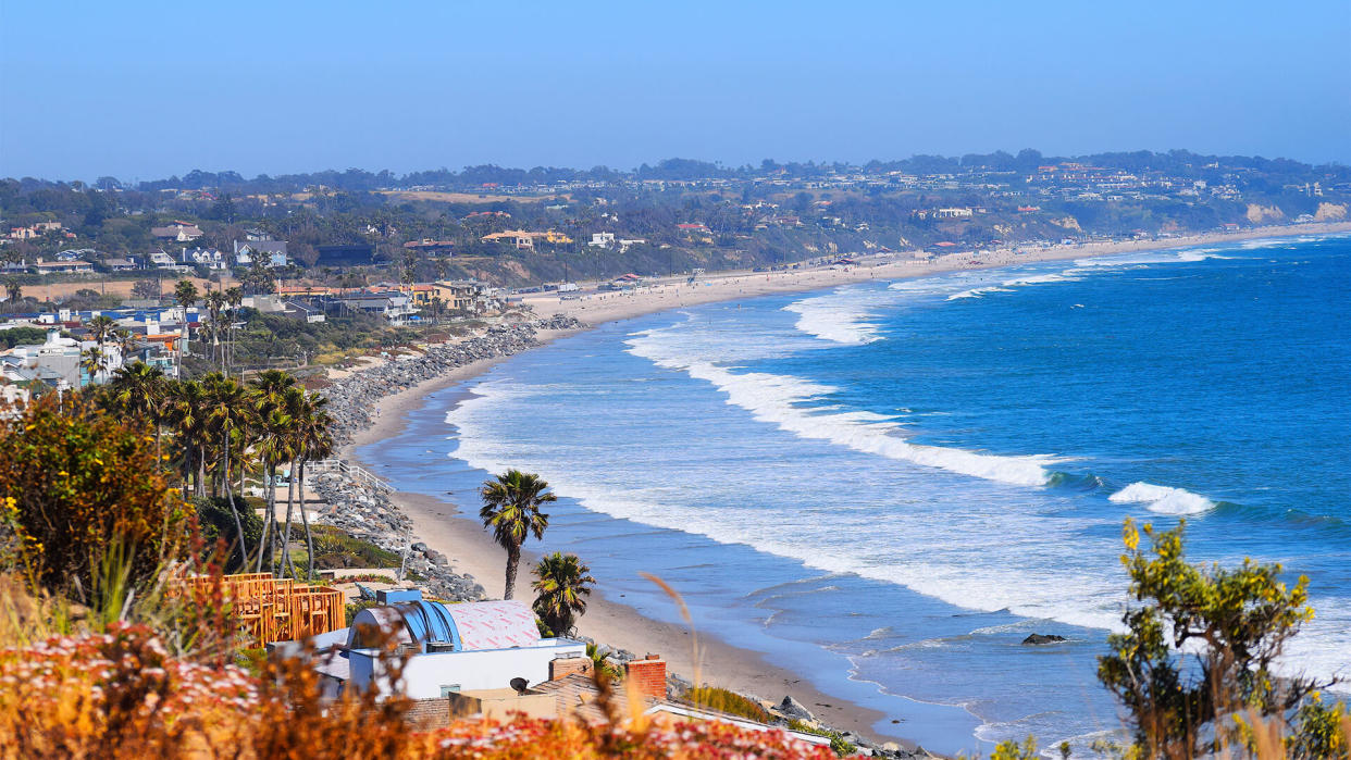 Malibu Beach coastline in California with the blue Pacific Ocean with waves coming in and beach with houses and palm tree's in background - Image.