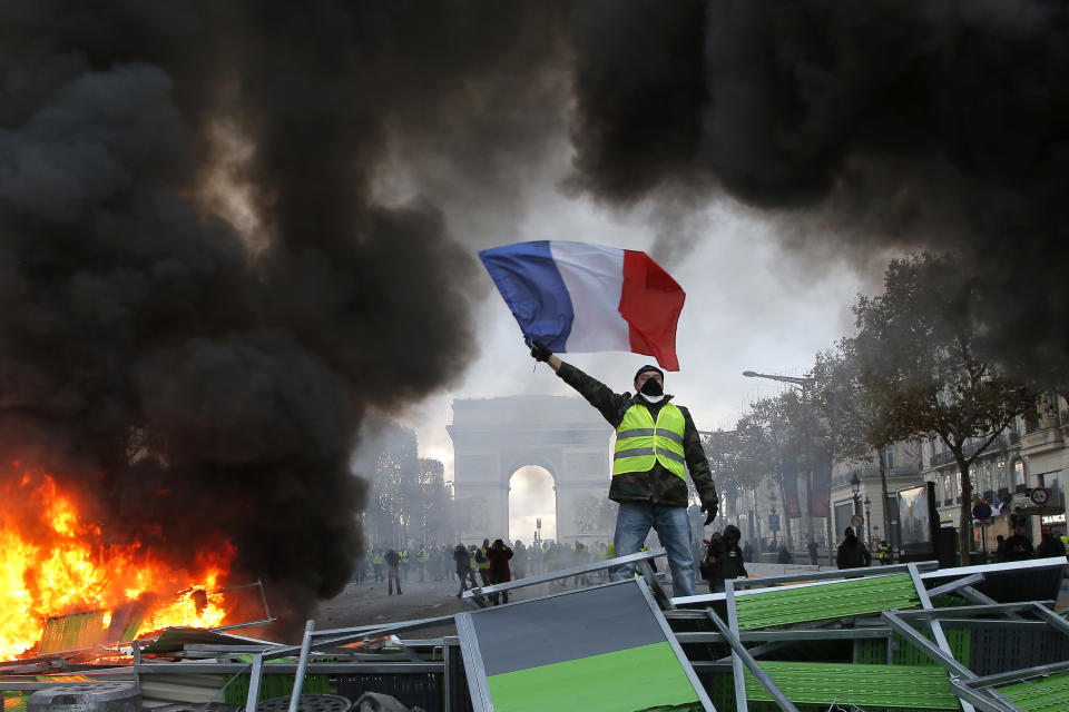 FILE - In this Nov. 24, 2018 file photo, a demonstrator waves the French flag on a burning barricade on the Champs-Elysees avenue with the Arc de Triomphe in background, during a demonstration against the rise of fuel taxes. Across the world, people are questioning truths they had long held to be self-evident, and they are dismissing some of them as fake news. They are replacing traditions they had long seen as immutable with haphazard reinvention. In France, people who feel left behind by a globalizing world have spent the last few weeks marching and rioting to protest a government they call elitist and out of touch. (AP Photo/Michel Euler, File)