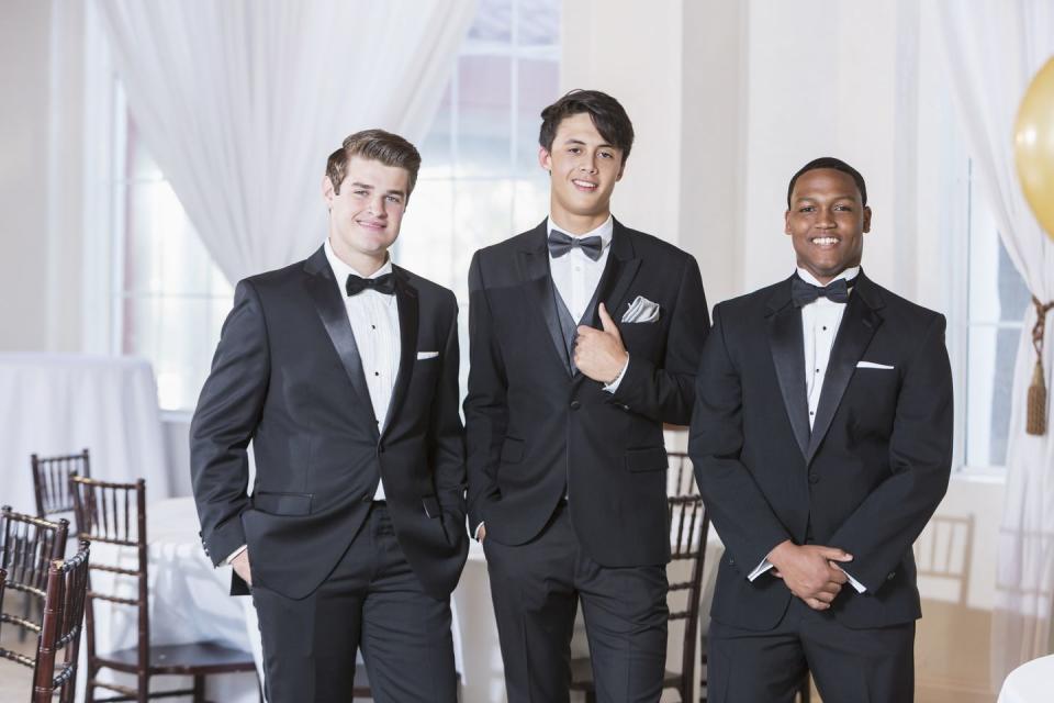 a group of three multi ethnic young men wearing tuxedos, standing in a row, smiling at the camera they are at a party, prom, a wedding or some other special formal event